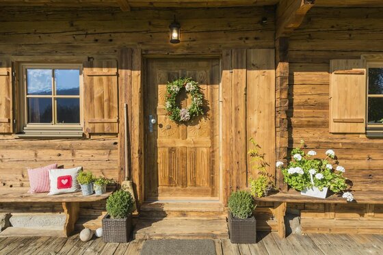 Eingangsbereich einer Holzhütte mit verzierter Tür, Türkranz, Blumenkästen und einer Holzbank mit Kissen.