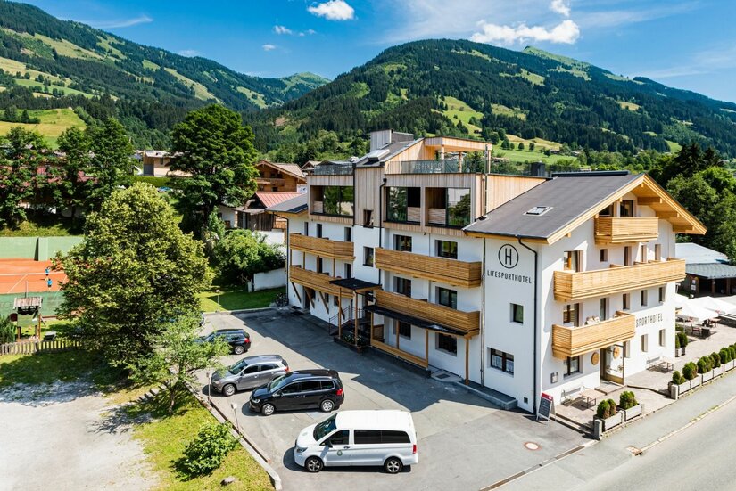 Hotel Hechenmoos in Berglandschaft Mehrstöckiges Hotelgebäude mit Holzbalkonen vor bewaldeten Bergen und einem Parkplatz im Vordergrund.