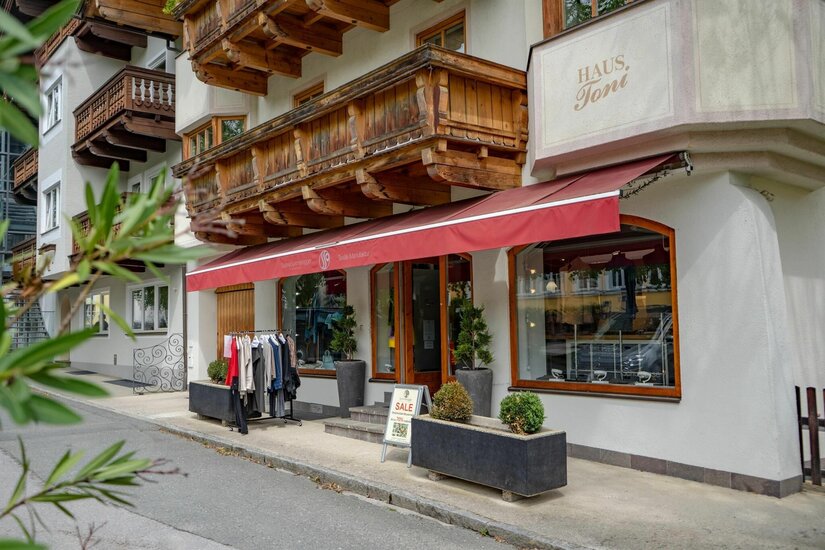 Sabine Sommeregger Manufaktur Eingangsbereich eines Bekleidungsgeschäfts mit roter Markise und Holzbalkon darüber.
