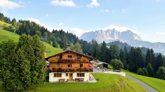 Wandhof vor dem Wilden Kaiser Traditionelles Holzhaus mit Blumenschmuck vor bewaldeten Hügeln und dem Wilden Kaiser Gebirge.