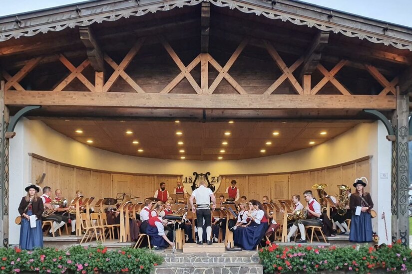 Musikpavillon Eine festliche Veranstaltung mit einer Musikgruppe auf einer Bühne. Die Musiker tragen traditionelle Kleidung und umgeben sich mit Stühlen und Blumen.
