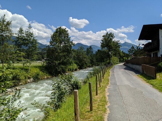 Asphaltierter Weg neben einem fließenden Fluss mit Bergen im Hintergrund.