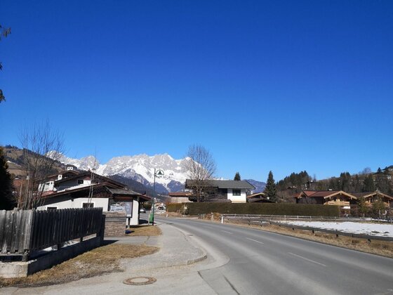 Siedlungsstraße mit Bergpanorama Asphaltierte Straße in einer Siedlung mit Blick auf schneebedeckte Berggipfel unter blauem Himmel.