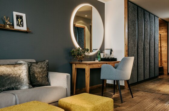 Zimmer im Grand Tirolia Modernes Hotelzimmer mit Sofa, Holztisch, beleuchtetem Rundspiegel und grauem Sessel.