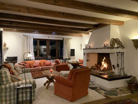 WZ1 Ein gemütliches Wohnzimmer mit einem offenen Kamin und rustikalen Holzbalken. Die Einrichtung besteht aus bequemen Sofas und einem warmen Ambiente.
