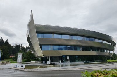 Reisebüro Eurotours Ein modernes, futuristisch gestaltetes Gebäude mit großen Fenstern und einer geschwungenen Fassadenstruktur. Es liegt in einer grünen Umgebung unter einem bewölkten Himmel.