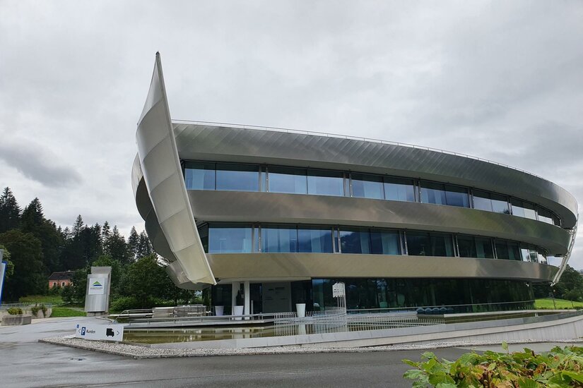 Ein modernes, futuristisch gestaltetes Gebäude mit großen Fenstern und einer geschwungenen Fassadenstruktur. Es liegt in einer grünen Umgebung unter einem bewölkten Himmel.