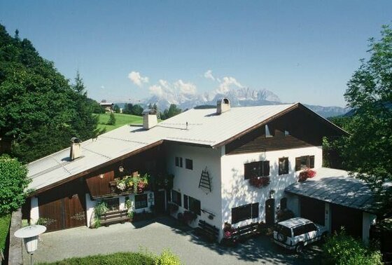 Landhaus Eva im Sommer Ein weißes Landhaus mit Holzelementen und Blumenschmuck vor einer Bergkulisse.