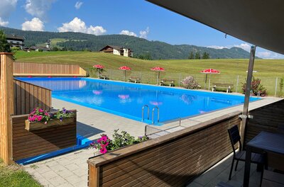 Ein schwimmendes, klares blaues Wasserbecken mit Blumen und Liegestühlen. Im Hintergrund sind grüne Wiesen und Berge sichtbar.