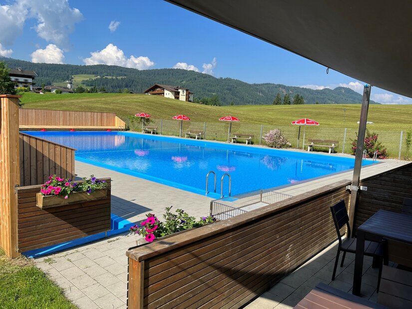 Ein schwimmendes, klares blaues Wasserbecken mit Blumen und Liegestühlen. Im Hintergrund sind grüne Wiesen und Berge sichtbar.