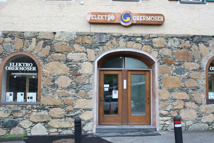 Obermoser Ein Gebäudeeingang mit zwei Holz-Türen und einem Schild mit der Aufschrift "Elektro Obermoser". Die Fassade ist aus Steinen und weist zwei Fenster auf.