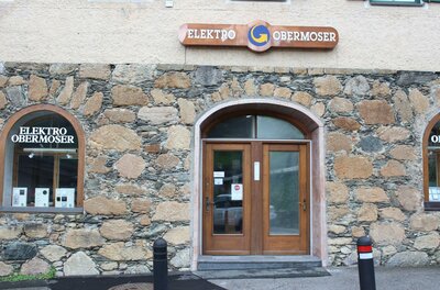 Obermoser Ein Gebäudeeingang mit zwei Holz-Türen und einem Schild mit der Aufschrift "Elektro Obermoser". Die Fassade ist aus Steinen und weist zwei Fenster auf.