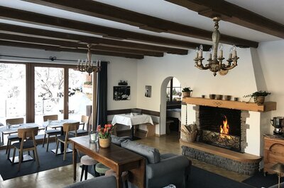 Innenansicht eines Gastraums mit brennendem Kamin, Holzbalkendecke und eingedeckten Tischen.