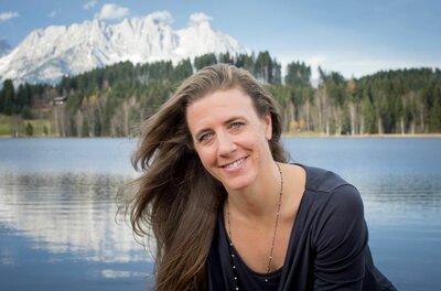 Porträt einer Frau vor einem Bergsee mit schneebedecktem Gebirgsmassiv im Hintergrund.