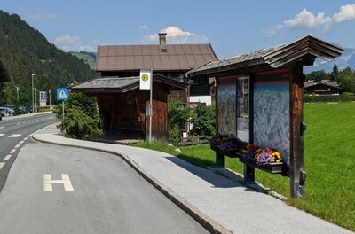 Bushaltestelle mit Holzunterstand und Informationstafel an einer Straße vor Bergkulisse.