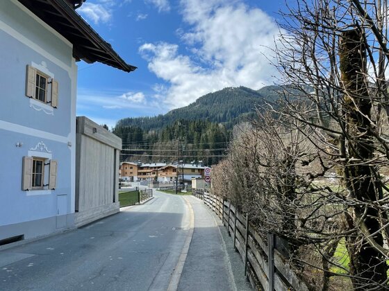 Auracher Höhenweg im Ort Asphaltierte Straße zwischen einem hellblauen Haus und einem Holzzaun mit Blick auf bewaldete Berge.