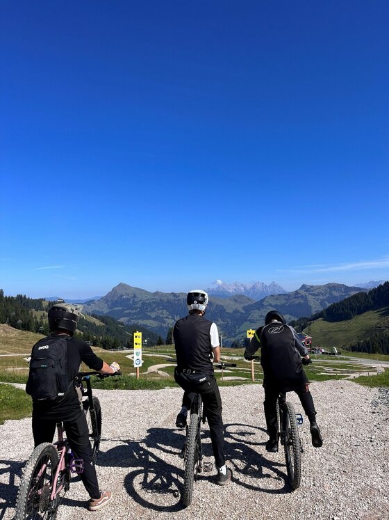 Drei Mountainbiker stehen mit ihren Rädern auf einem Schotterweg vor einem Bergpanorama unter blauem Himmel.