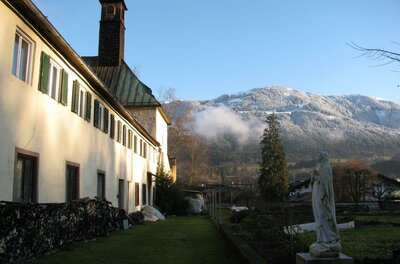 Ein charmantes Gebäude mit grünen Fensterläden und einem blauen Himmel im Hintergrund. Im Vordergrund steht eine Statue und die Umgebung ist von schneebedeckten Bergen umgeben.