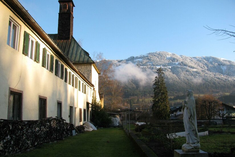 Kloster Ein charmantes Gebäude mit grünen Fensterläden und einem blauen Himmel im Hintergrund. Im Vordergrund steht eine Statue und die Umgebung ist von schneebedeckten Bergen umgeben.