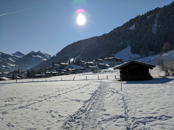 Winterlandschaft bei Malerndörfl Verschneite Wiese mit Spuren im Schnee, einer Holzhütte und einem Dorf vor bewaldeten Bergen unter blauem Himmel.