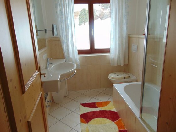 Badezimmer Apartment 1 Badezimmer mit Waschbecken, WC, Badewanne und Fenster mit Blick auf verschneite Bäume.