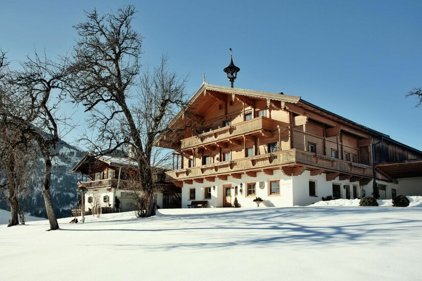 Alpiner Bauernhof aus Holz und Stein in einer verschneiten Winterlandschaft unter blauem Himmel.