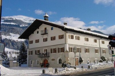 Traditionelles Gasthaus mit weißer Fassade und braunen Fensterläden im Schnee vor bewaldeten Bergen.