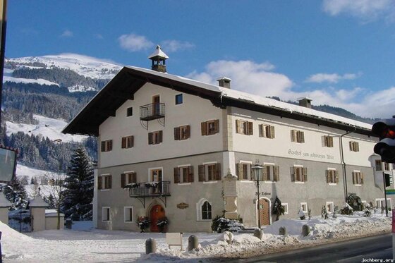 Gasthof Schwarzer Adler im Winter Traditionelles Gasthaus mit weißer Fassade und braunen Fensterläden im Schnee vor bewaldeten Bergen.