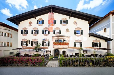 01Eggerwirt001 Ein charmantes Hotelgebäude mit traditioneller bayerischer Architektur. Es hat viele Fenster und eine einladende Terrasse im Freien.