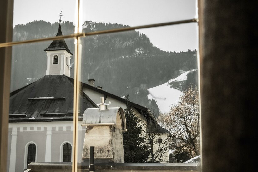 Apart_Mantinger_Kitz_Klammer-07736 Ein Blick aus einem Fenster auf ein malerisches Dorf mit einer Kirche. Im Hintergrund sind schneebedeckte Berge zu sehen.