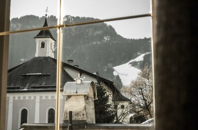 Ein Blick aus einem Fenster auf ein malerisches Dorf mit einer Kirche. Im Hintergrund sind schneebedeckte Berge zu sehen.