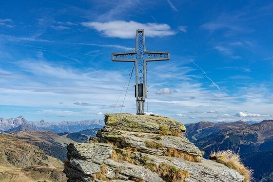 Ein metallisches Gipfelkreuz auf einem felsigen Berggipfel vor blauem Himmel und Gebirgspanorama.