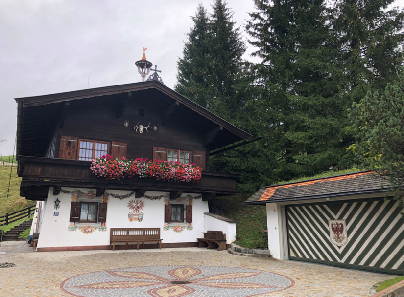 Traditionelles Alpenhaus mit dunkler Holzfassade, Blumenbalkon und Wandmalereien vor Nadelbäumen.