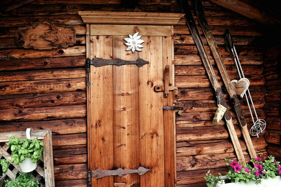 Holztür einer Almhütte mit Edelweiß-Verzierung, daneben lehnen alte Holzski an der Blockwand.