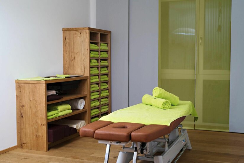 Eine braune Massageliege mit grünen Handtüchern vor einem Holzregal in einem hellen Behandlungsraum.