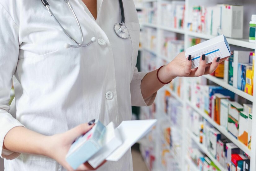 Person im weißen Kittel hält Medikamentenschachteln vor einem Apothekenregal.