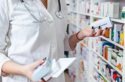 Person im weißen Kittel hält Medikamentenschachteln vor einem Apothekenregal.