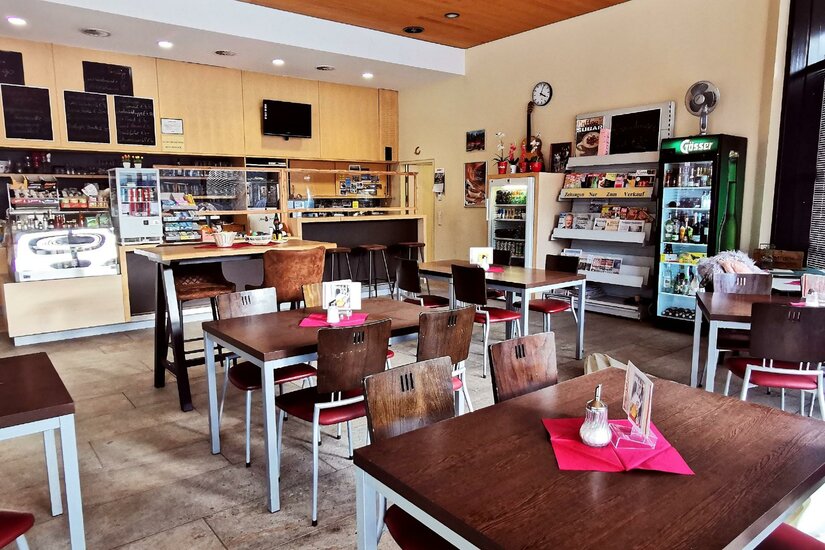 Bahnhofkaffee Ein gemütliches Café mit Holzstühlen und Tischen. Im Hintergrund sind Getränke und Snacks in einem Regal zu sehen.