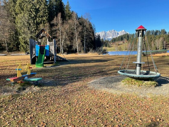 Spielplatz am Schwarzsee Kinderspielplatz auf einer Wiese mit Rutsche, Wippe und Karussell vor einem See und Bergen.