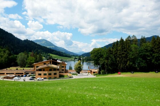 Hörpfing Runde am See Blick über eine grüne Wiese auf ein Holzgebäude vor einem See und bewaldeten Bergen.
