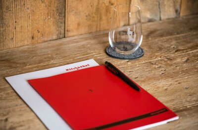 Ein rotes Notizbuch liegt auf einem Holztisch neben einem Glas und einem Stift. Der Tisch hat eine rustikale, natürliche Oberfläche.