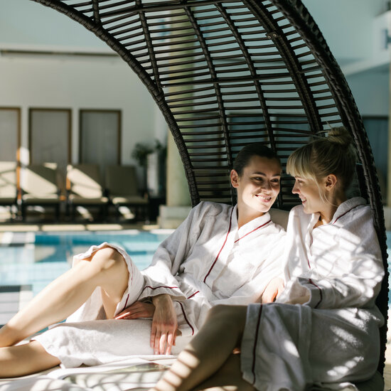 Zwei Frauen in Bademänteln sitzen entspannt neben einem Pool und genießen einen Wellnesstag in Kitzbühel.