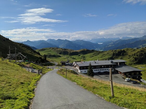 Asphaltierter Wanderweg führt bergab zu einem großen Berggasthof in einer alpinen Wiesenlandschaft mit Bergpanorama.