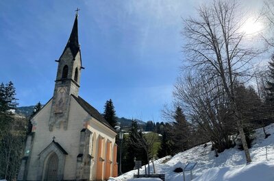 Kleine Kapelle mit spitzem Turm in einer verschneiten Winterlandschaft unter blauem Himmel.