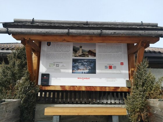 Informationstafel aus Holz mit Dach und Sitzbank vor Nadelgehölzen.