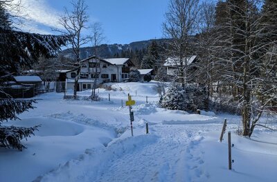 Verschneiter Wanderweg führt zu einem gelben Wegweiser vor alpinen Häusern und Nadelbäumen.