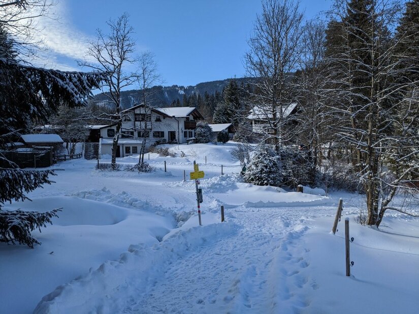 Verschneiter Wanderweg führt zu einem gelben Wegweiser vor alpinen Häusern und Nadelbäumen.
