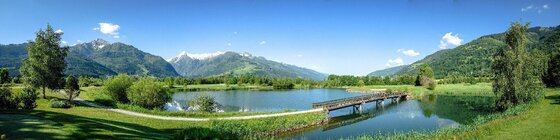 Golfclub Zell am See Panorama eines Sees mit Holzbrücke, umgeben von Wiesen und Bergen unter blauem Himmel.