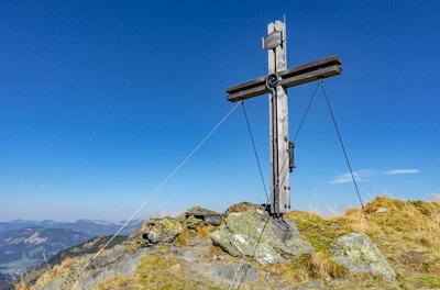 Ein hölzernes Gipfelkreuz auf einem felsigen Berggipfel vor blauem Himmel.