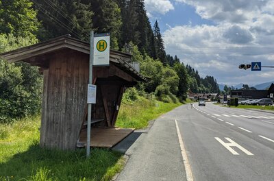 Hölzernes Wartehäuschen einer Bushaltestelle an einer Landstraße neben einem Wald.